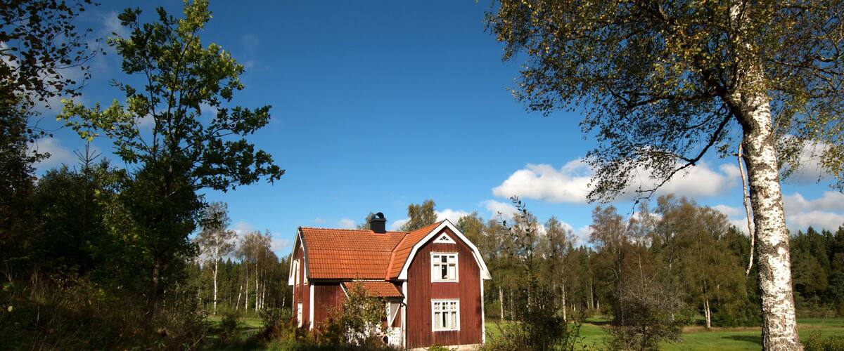 A red wooden house with a white trim, surrounded by trees and a grassy field.