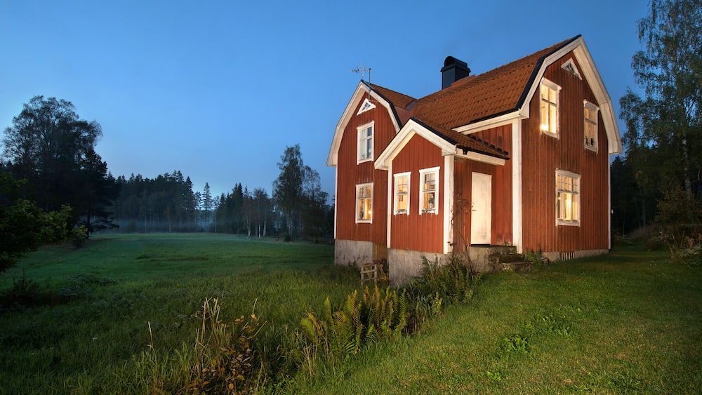 Ein rotes Holzhaus mit geneigtem Dach und weißem Zierdekor, umgeben von einer grünen Wiese und Bäumen.