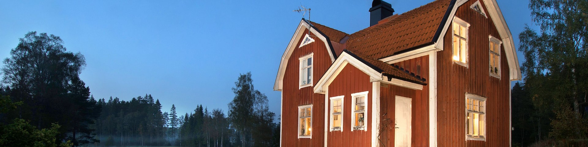 A red wooden house with a sloped roof and white trim, surrounded by a grassy field and trees.
