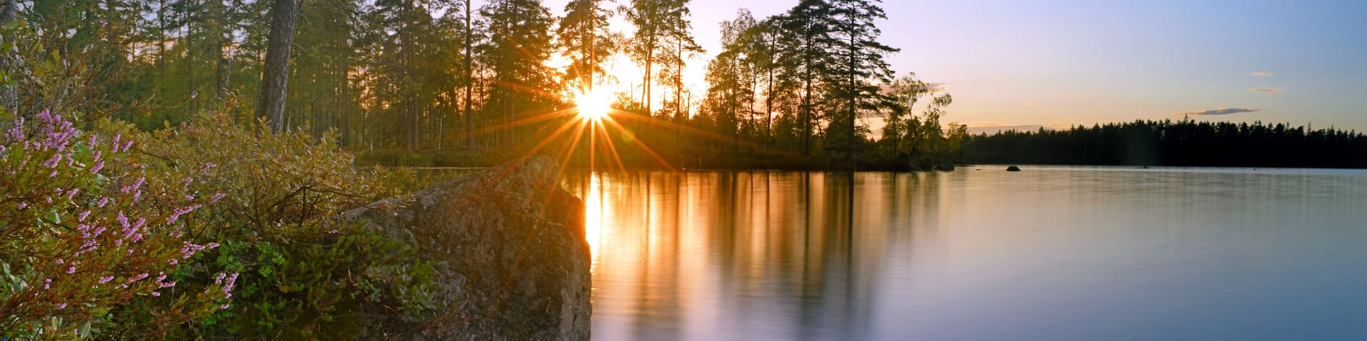 A serene lake at sunset with a rocky shore and dense trees in the background.