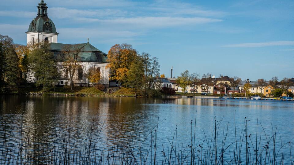 A historic church with a green dome and a bell tower, surrounded by autumn trees and a calm lake.