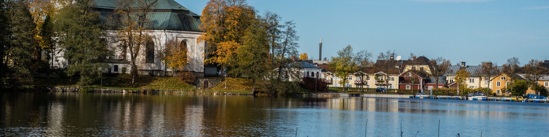 A historic church with a green dome and a bell tower, surrounded by autumn trees and a calm lake.