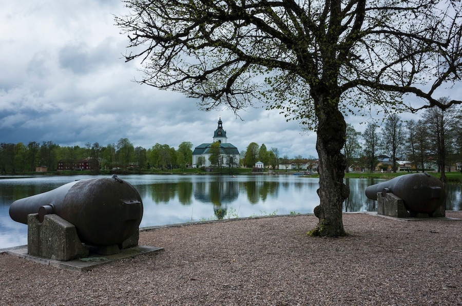 Filipstad's Church and cannons on Kanonudden in Filipstad, White church building in the distance is reflecting in the water, Two large cannons, A dark tree with small leaves, Cloudy weather