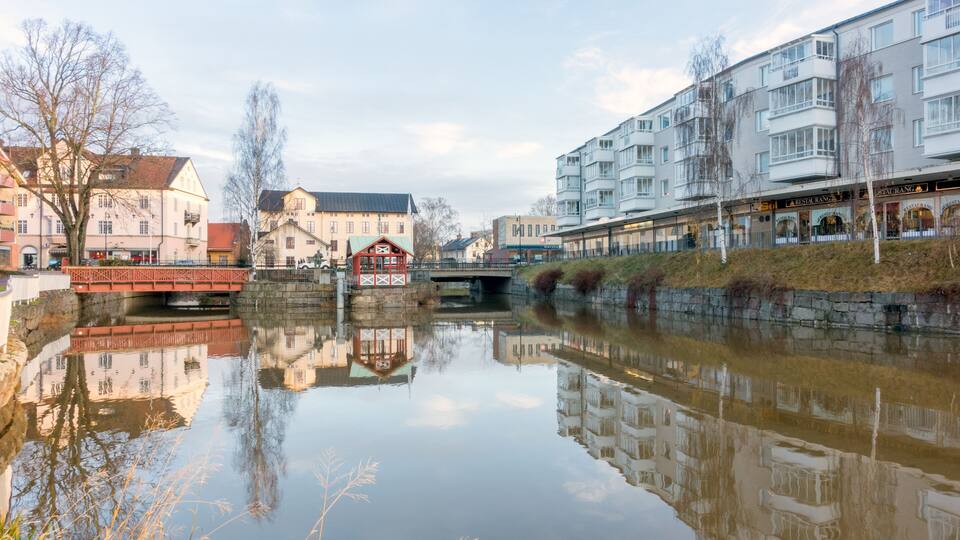 A calm river reflecting buildings, a red bridge, and a stone wall.