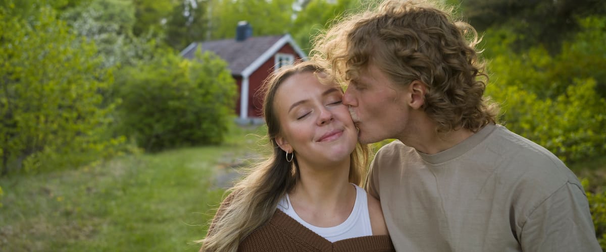 A couple embracing in a lush green outdoor setting with a red house in the background.