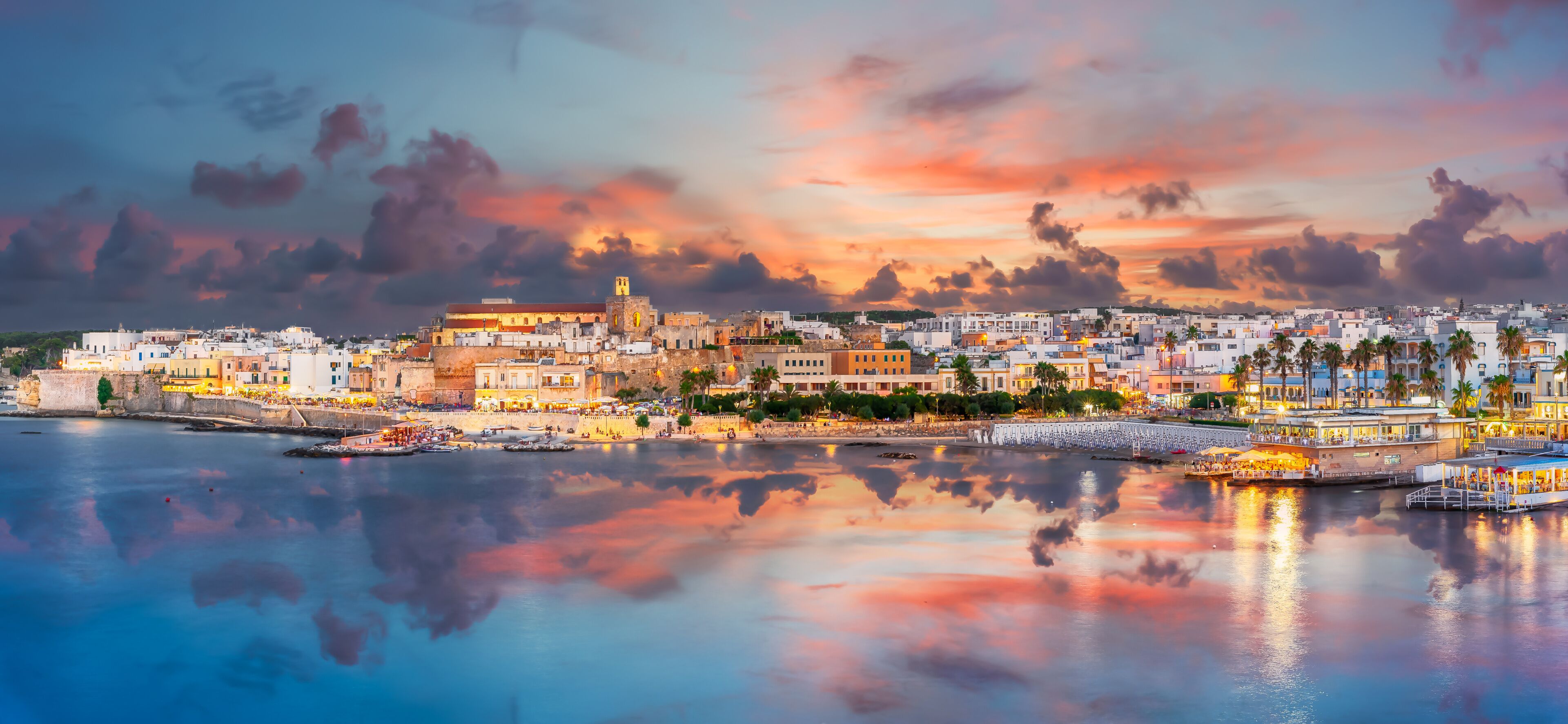 A coastal cityscape at sunset with a prominent church, waterfront promenade, and a reflection of the sky in the calm sea.