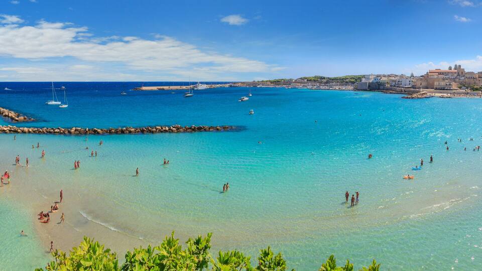 A coastal town with clear turquoise waters, a rocky breakwater, and buildings along the shore.