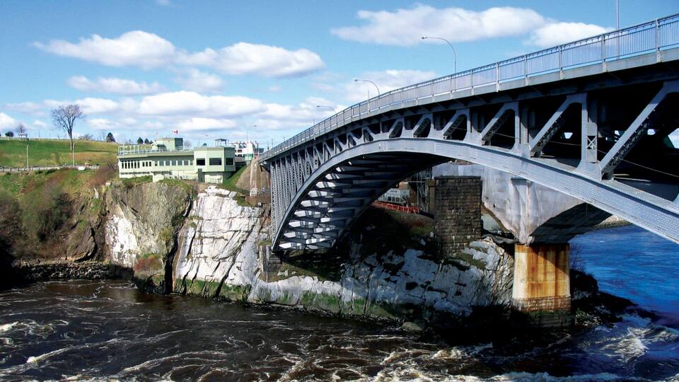 Un pont d’acier enjambant une rivière, avec des falaises rocheuses et des bâtiments en arrière-plan.