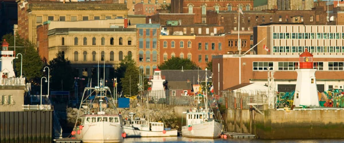 A harbor with several boats docked, a lighthouse, and historic buildings in the background.