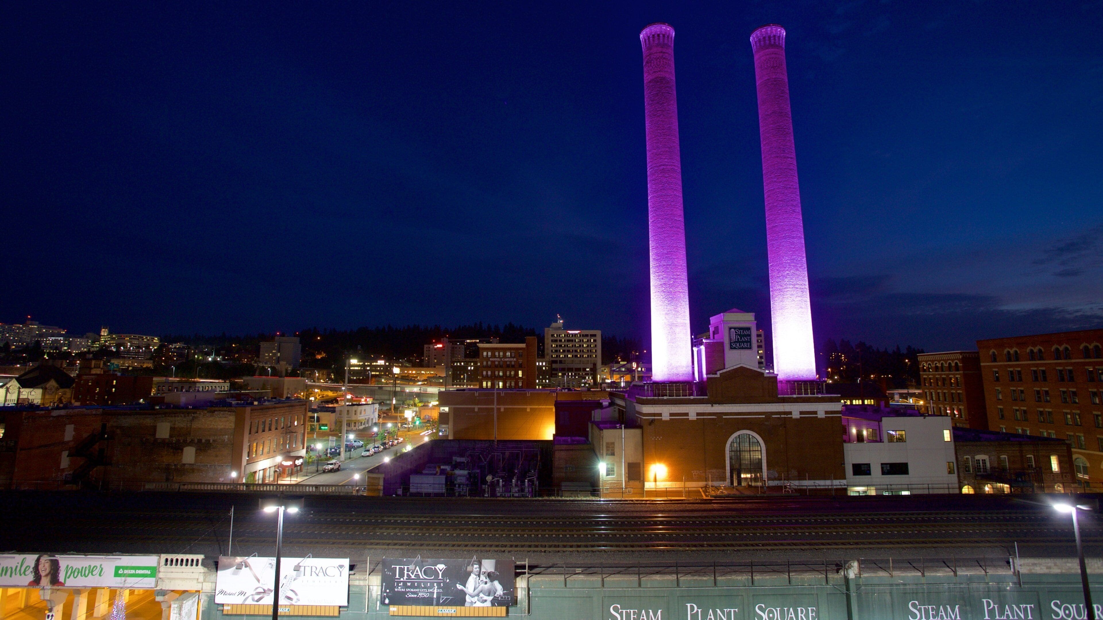 A cityscape at night with prominent purple-lit smokestacks of a plant facility.