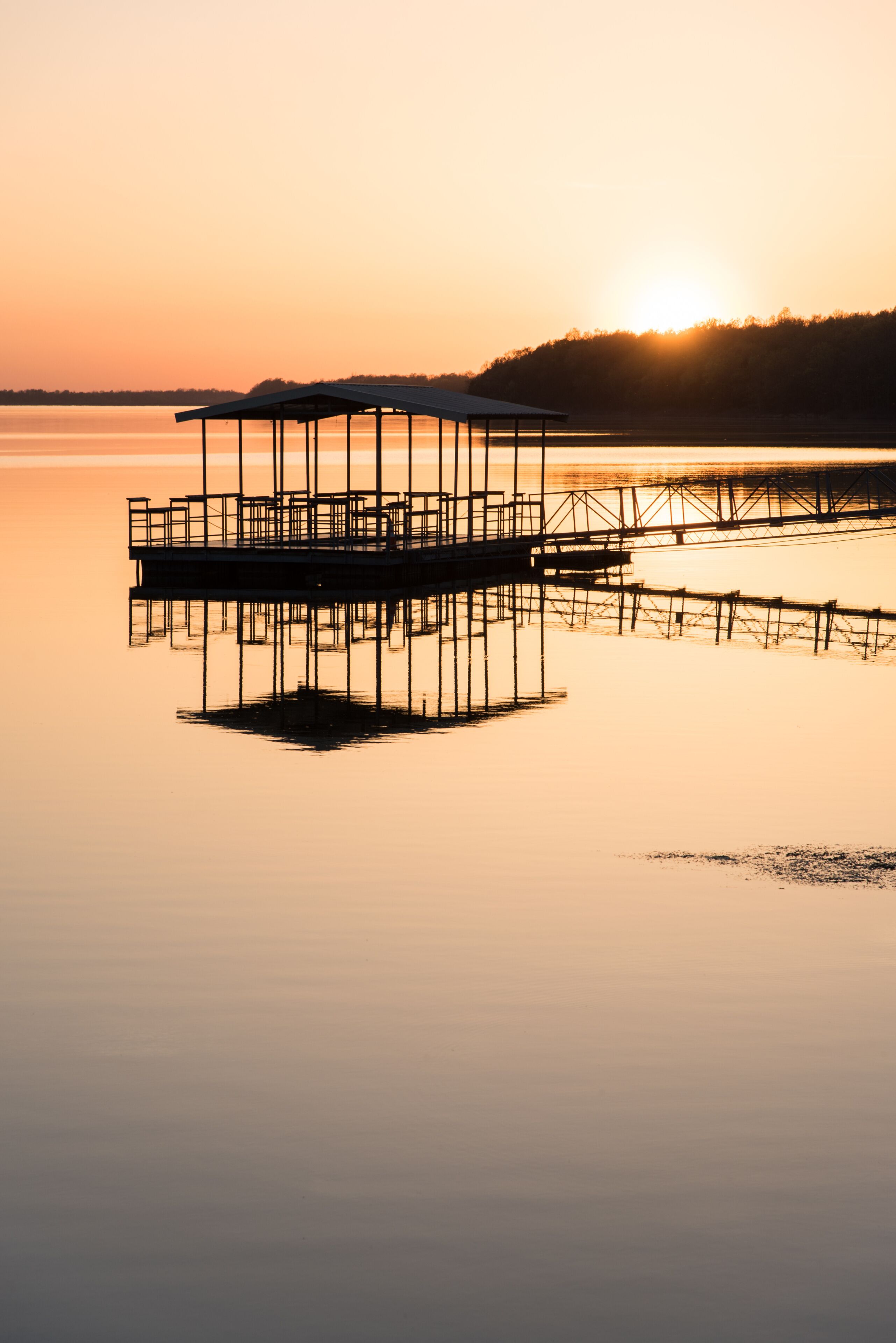 A pier extending into calm waters at sunset with a forested hill in the background.