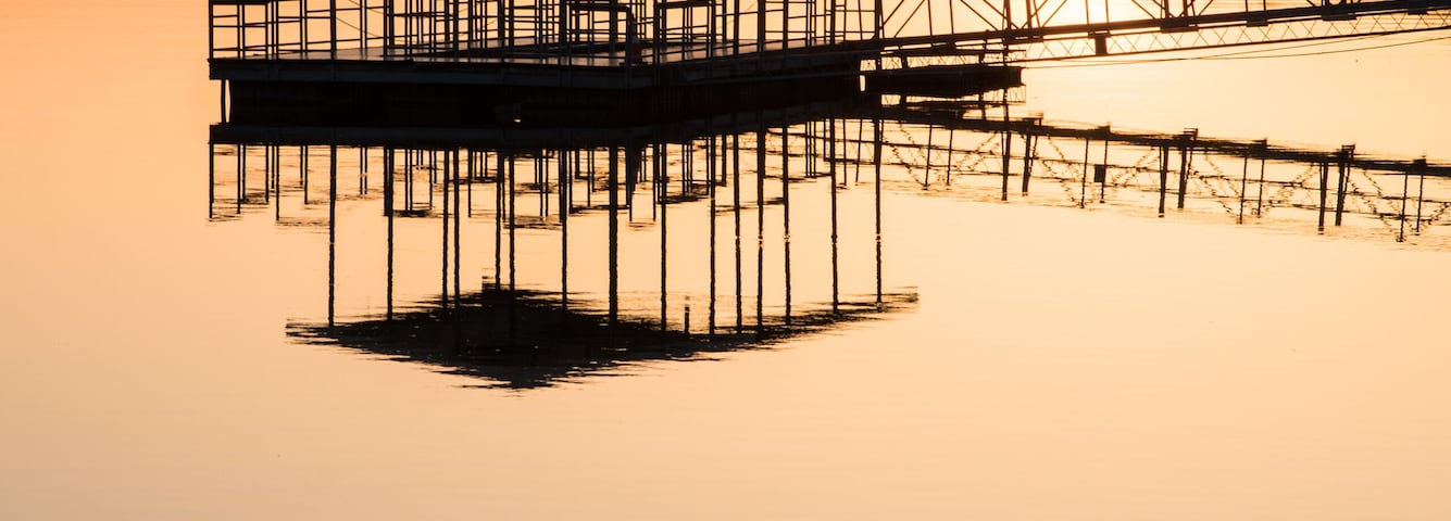 Un muelle se adentra en aguas tranquilas al atardecer, con una colina boscosa en el fondo.