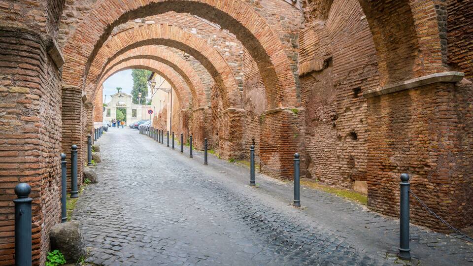An arched brick pathway with cobblestone pavement, flanked by brick walls and black bollards.