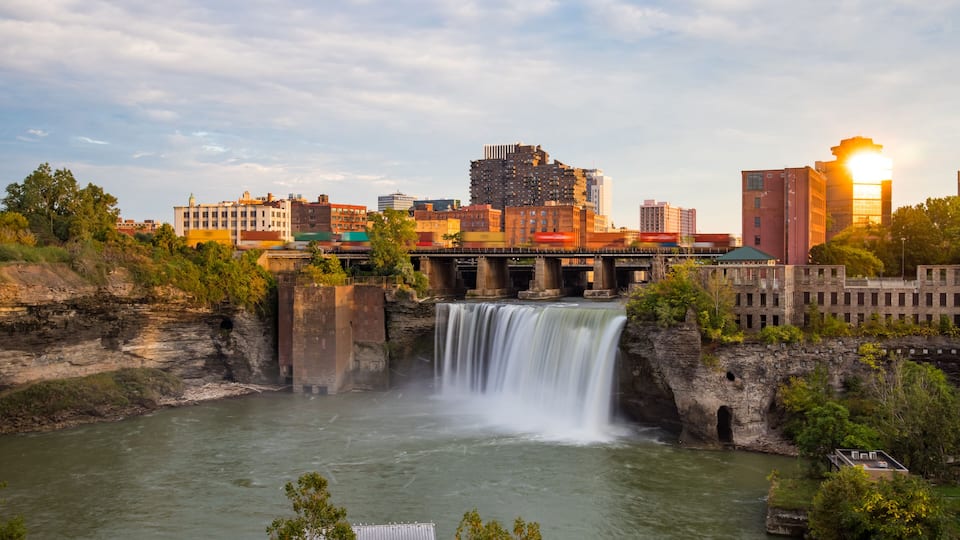 A waterfall cascading into a river, surrounded by urban buildings and greenery.