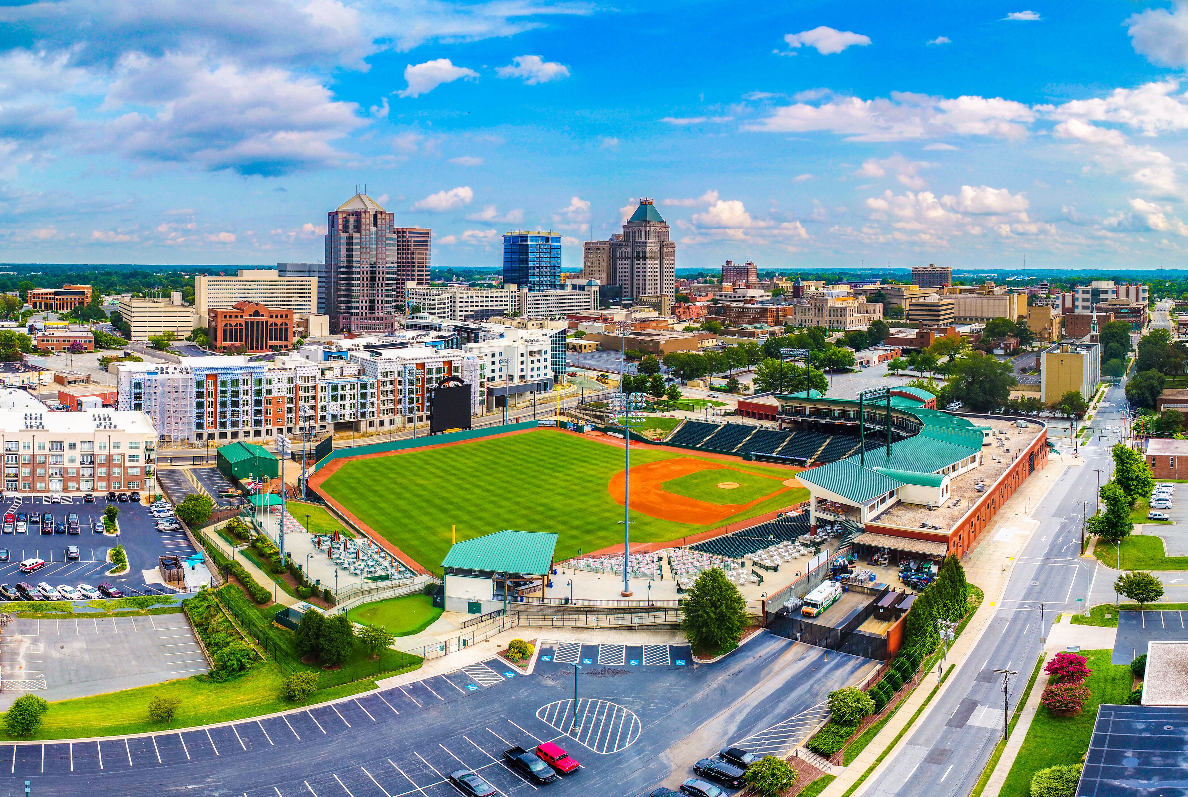 A cityscape with a baseball stadium surrounded by urban buildings and a parking lot.