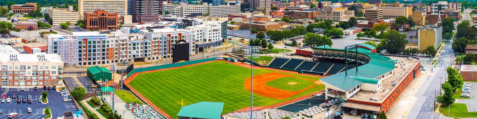 A cityscape with a baseball stadium surrounded by urban buildings and a parking lot.