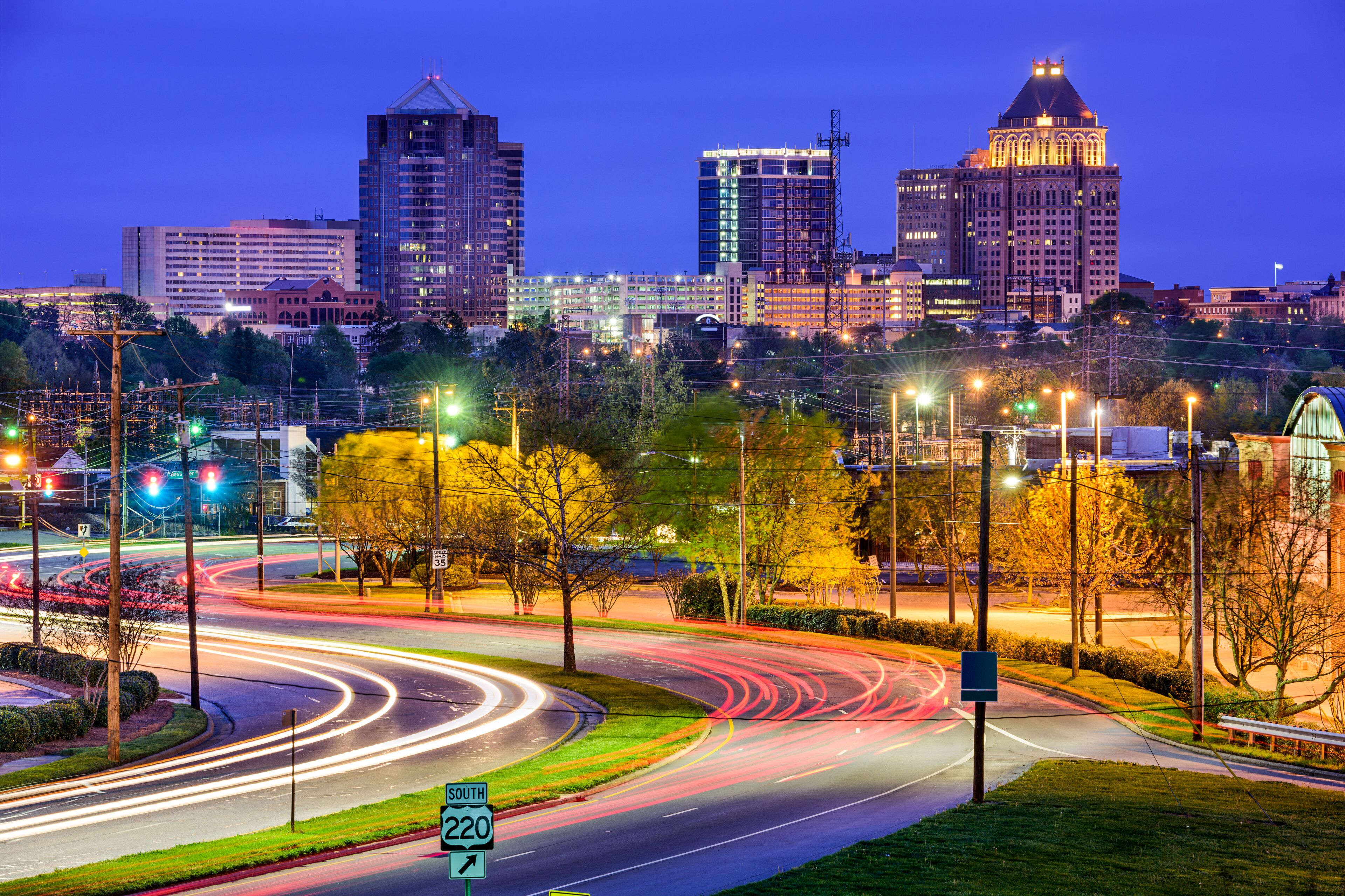 A cityscape at dusk with illuminated buildings, a roundabout, and a sign for route 220.