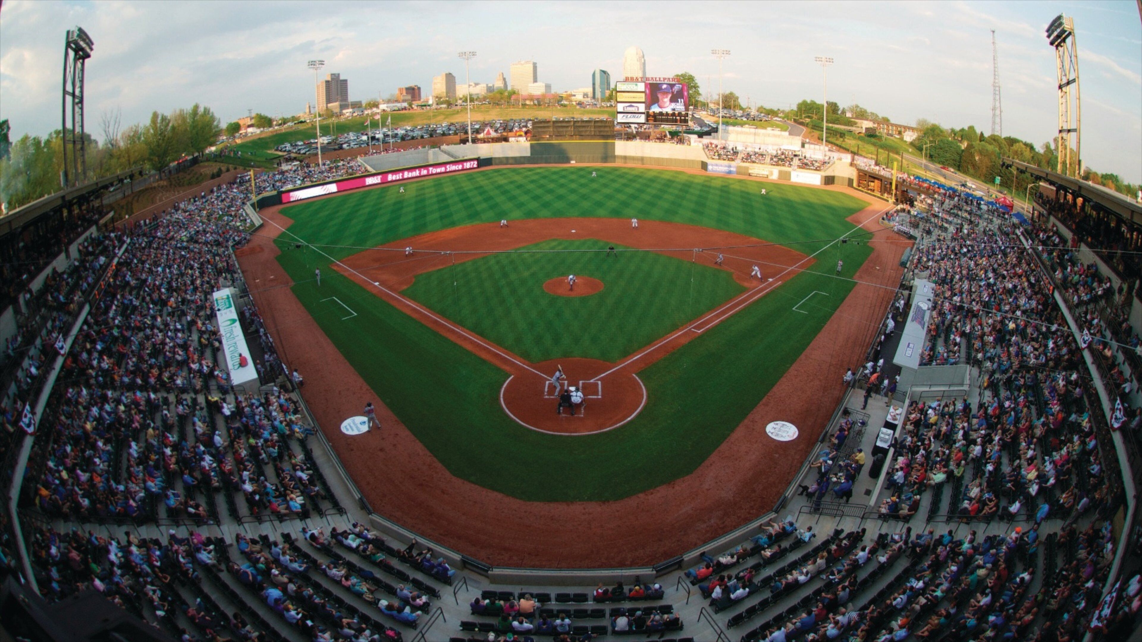 A baseball stadium with a large crowd, a baseball diamond, and city buildings in the background.