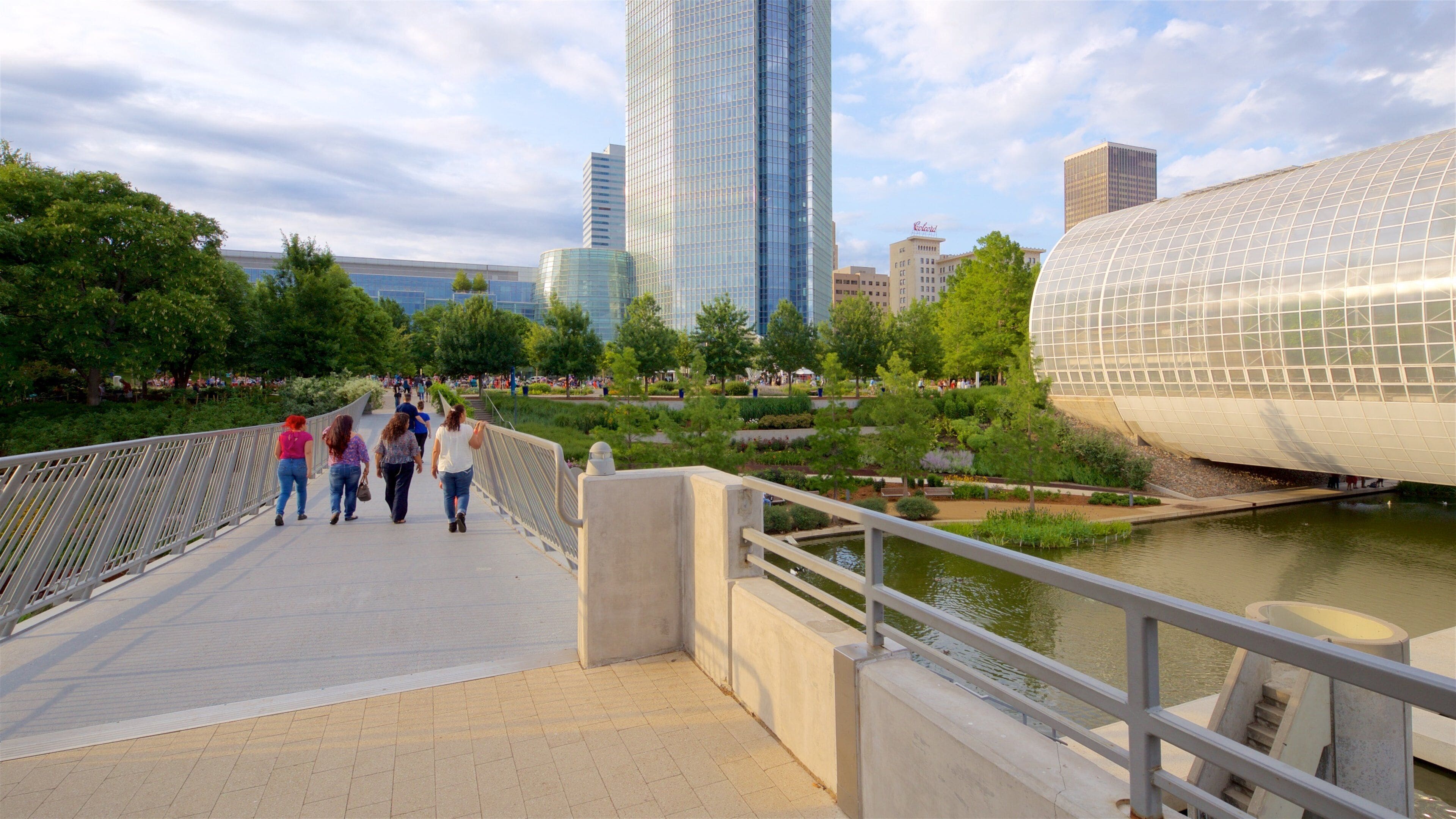 A pedestrian bridge with a river, modern buildings, and greenery.