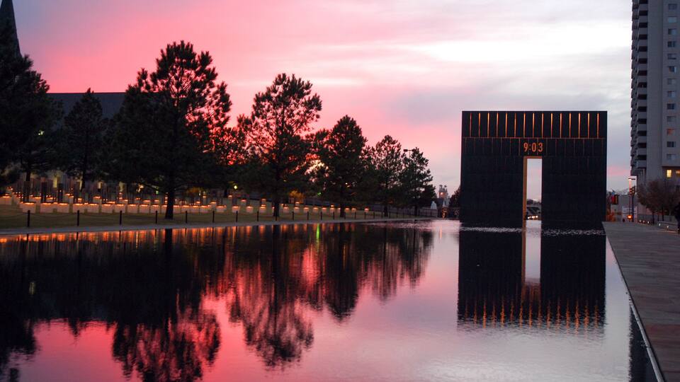 A sunset over a reflective water body with trees and a lit-up archway displaying the time.
