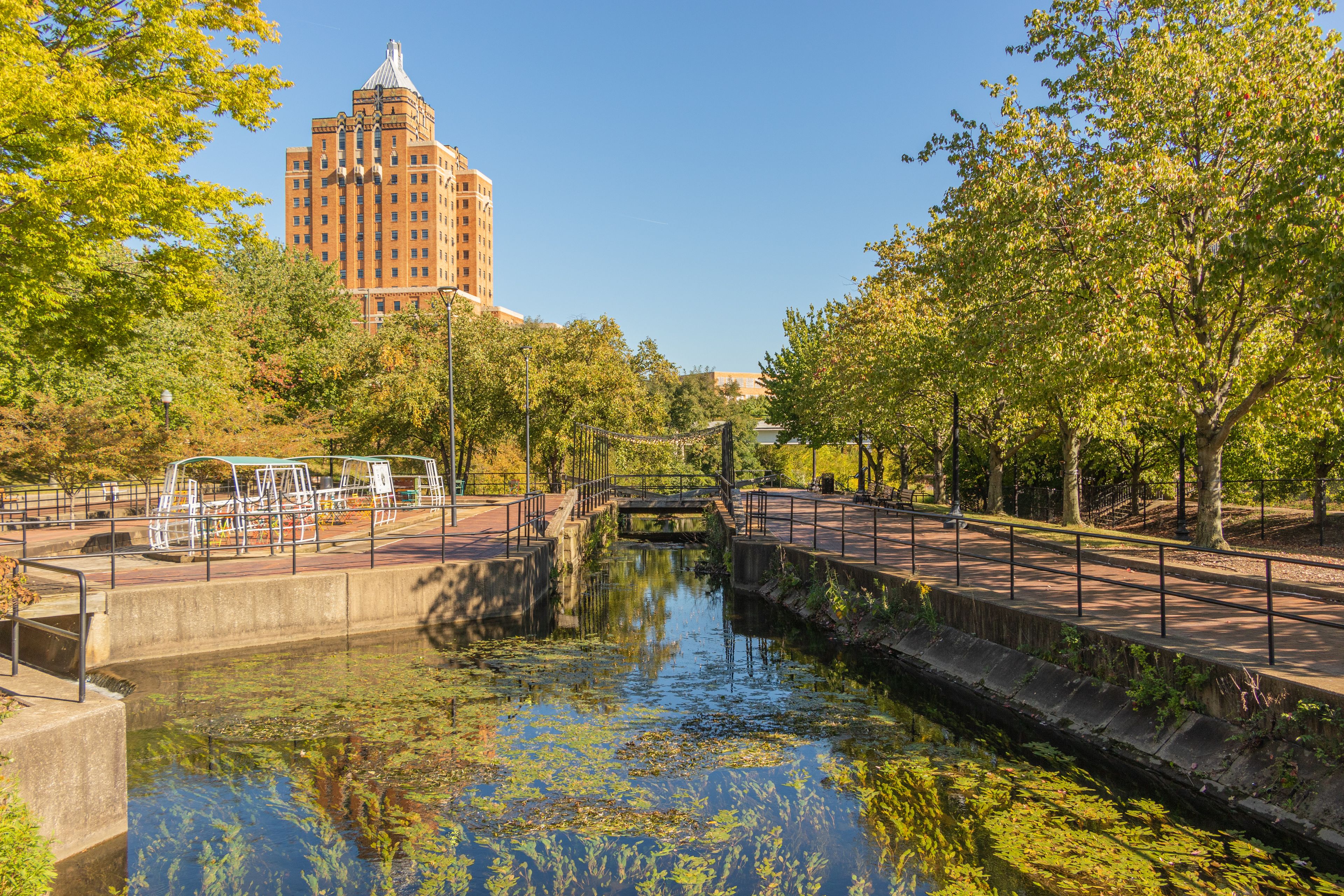 A canal with a bridge, a park with trees, and a large building in the background.