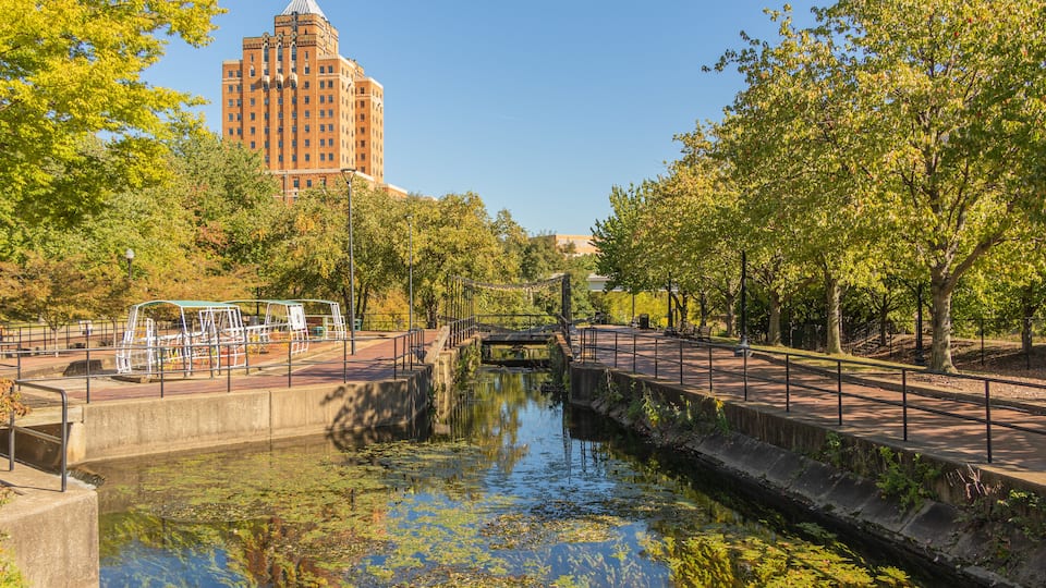 A canal with a bridge, a park with trees, and a large building in the background.