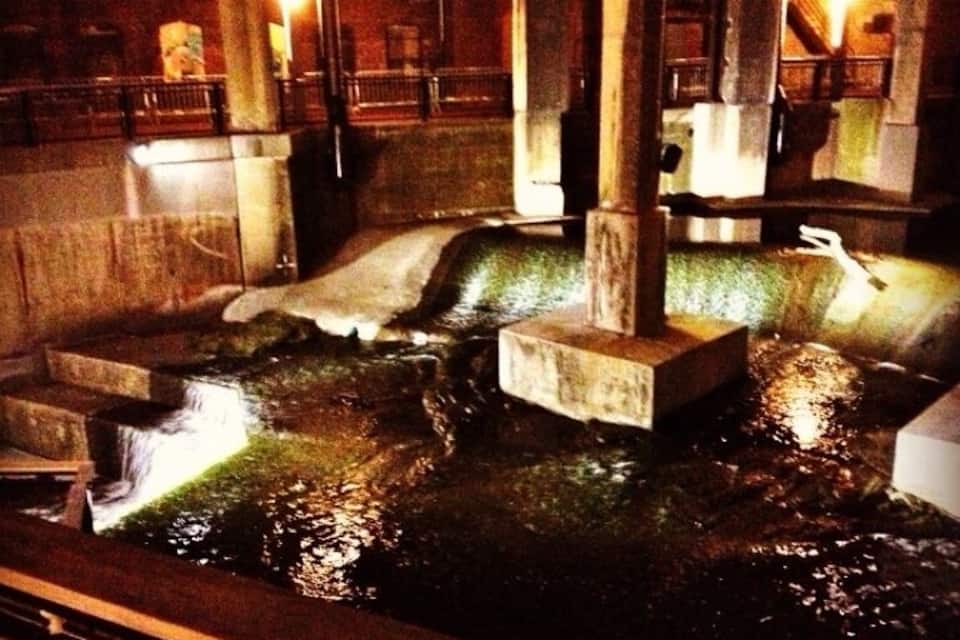 A nighttime view of a stone water feature with steps and a central column, surrounded by stone walls and illuminated by artificial lighting.