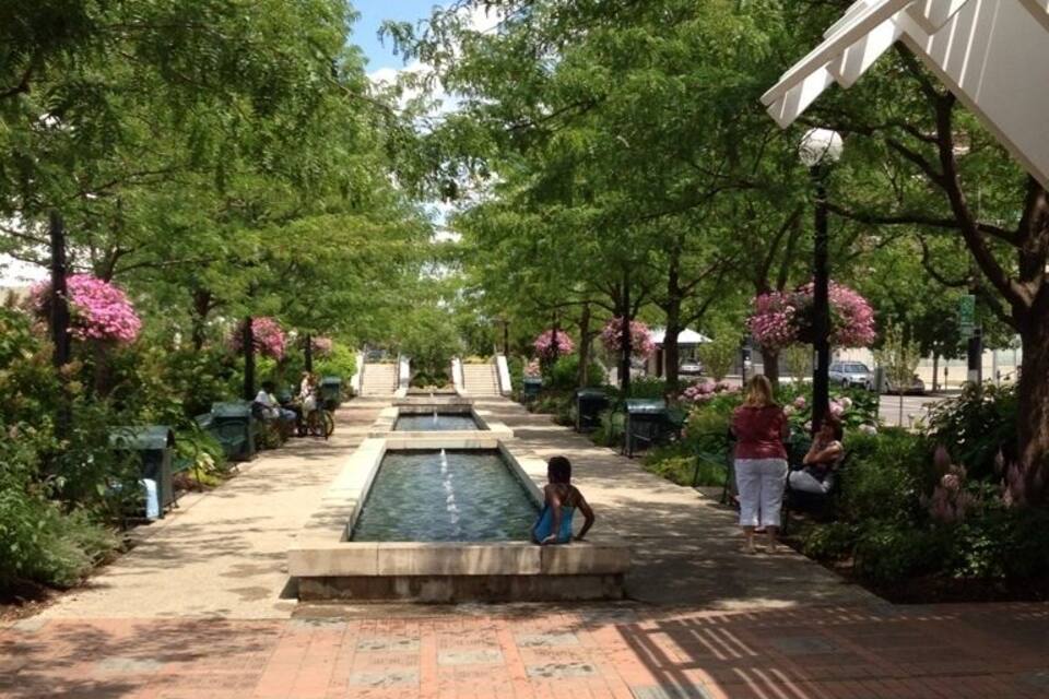 A paved pathway with a central water feature, surrounded by trees and benches.