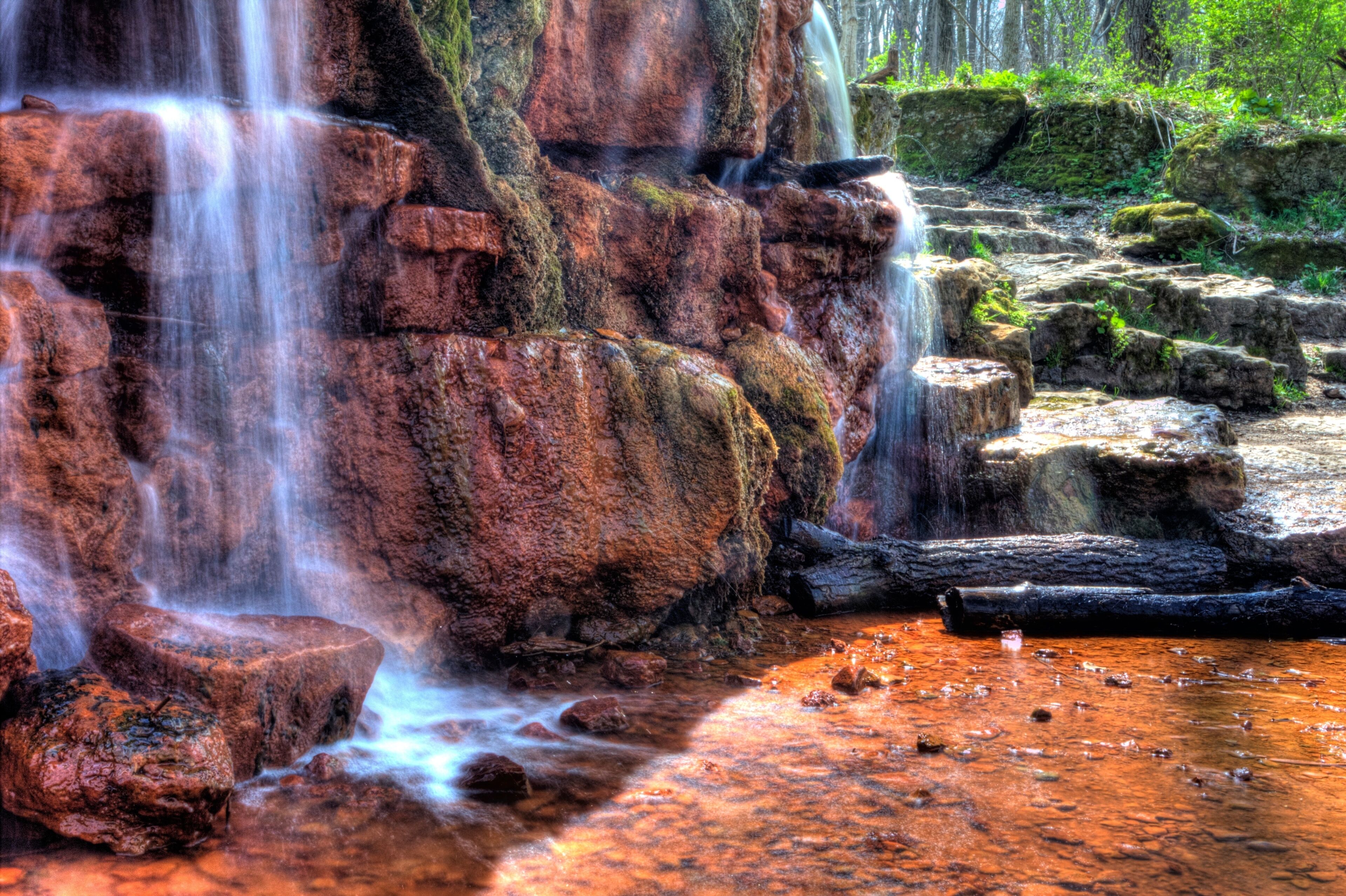 Una cascada fluye sobre rocas rojizo-marrón, con un estanque de agua en la base.