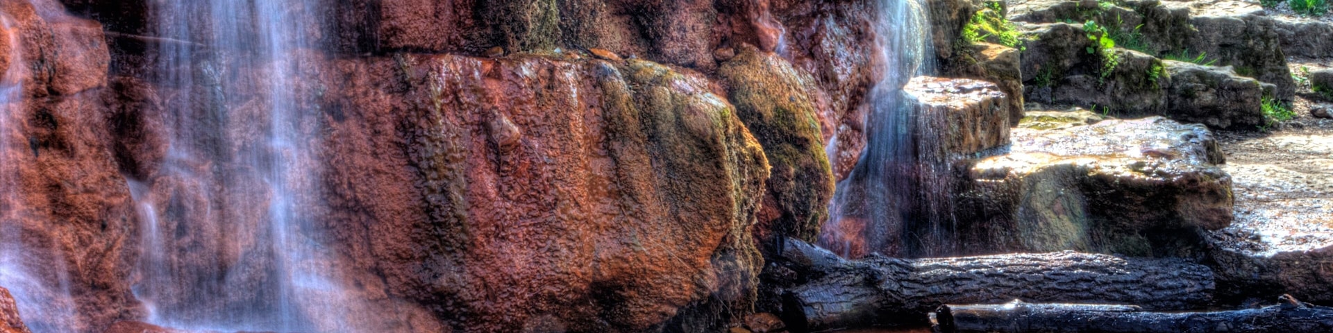 Una cascada fluye sobre rocas rojizo-marrón, con un estanque de agua en la base.