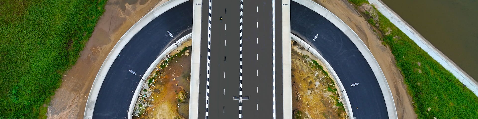 An aerial view of a curved road with a central divider, surrounded by greenery and a body of water.