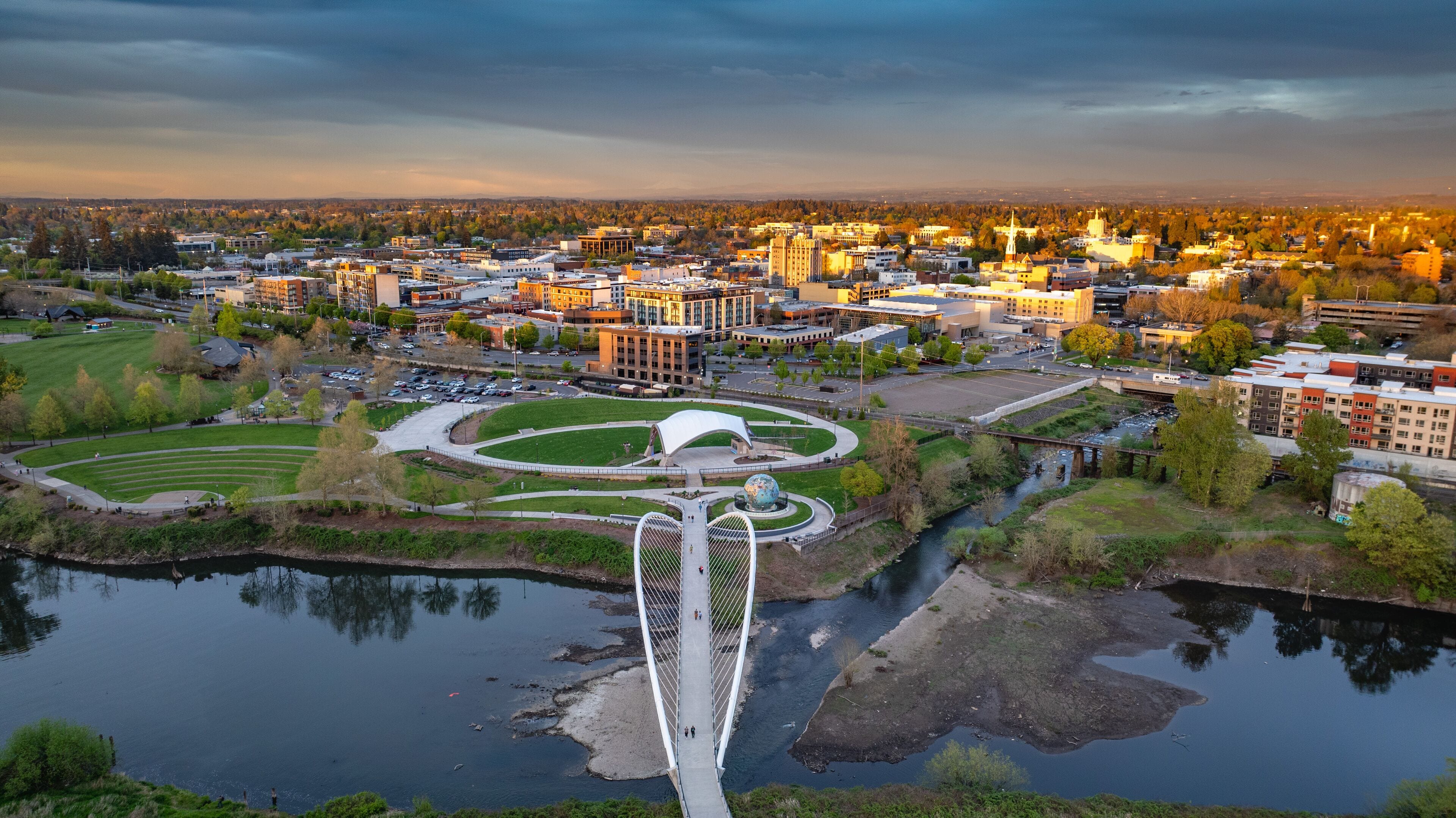 A cityscape with a prominent bridge over a river, surrounded by urban buildings and a park with a globe sculpture.
