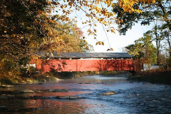 A red covered bridge over a river with autumn leaves.