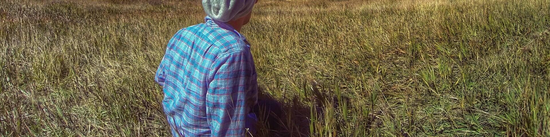 A person in a plaid shirt and gray hat sitting in a field with autumn-colored trees and mountains in the background.