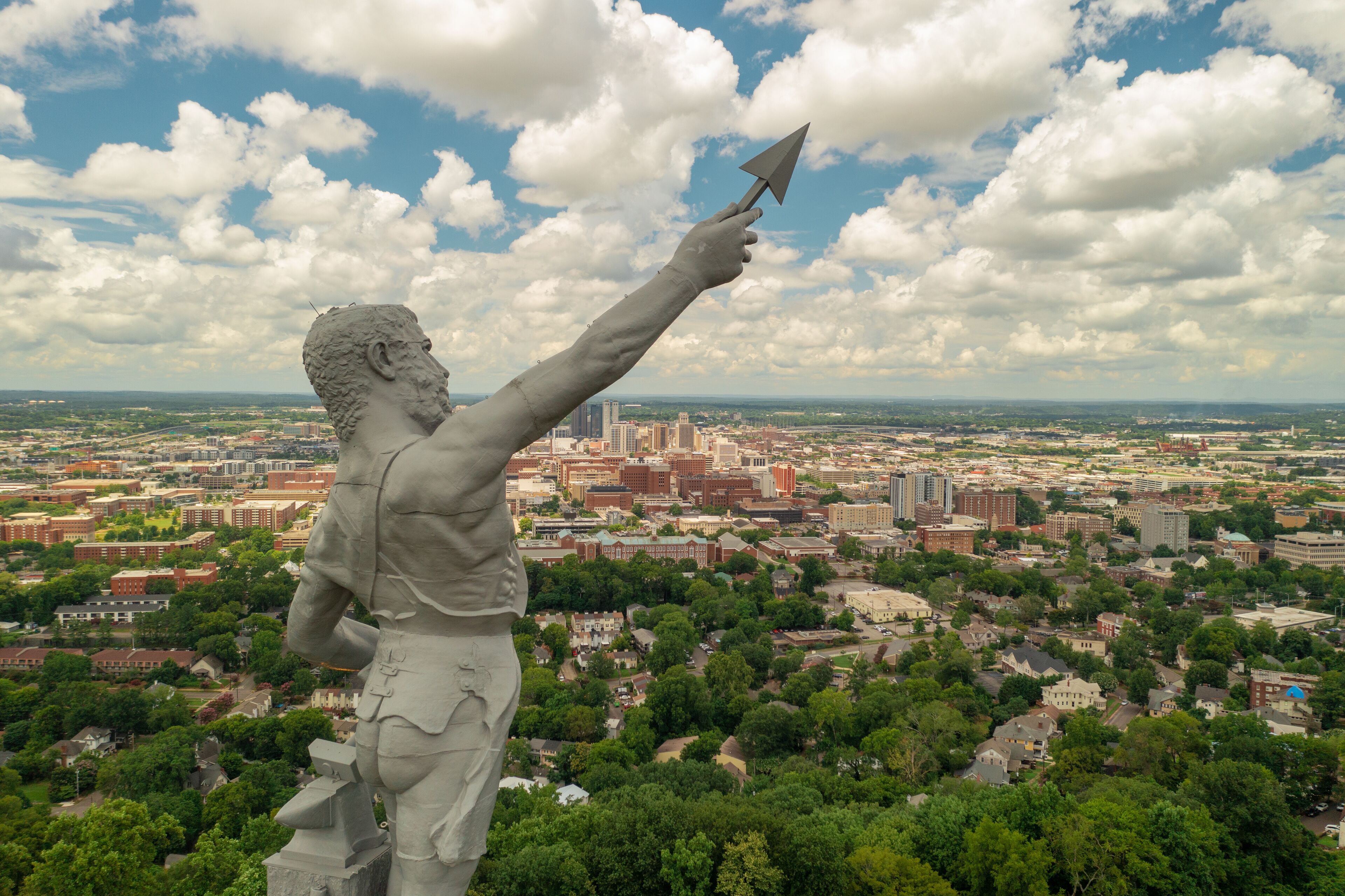 A statue of a figure holding a spear, overlooking a cityscape with buildings and greenery.