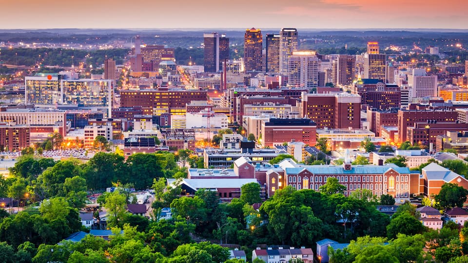 A cityscape at sunset with a mix of modern and historic buildings, dense urban layout, and surrounding greenery.