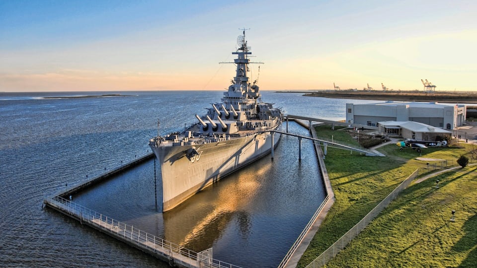 Un gran buque de guerra atracado en un muelle, con una zona verde y edificios industriales al fondo.