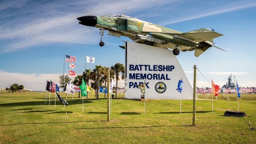 Un avión de combate volando bajo sobre un campo de hierba, con un cartel del Parque Conmemorativo del acorazado al fondo.