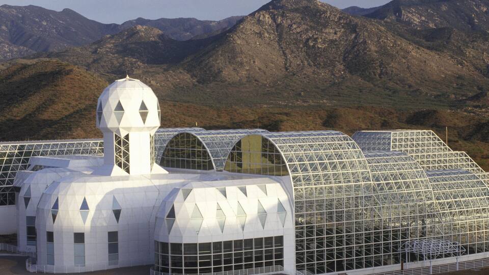 A modern greenhouse complex with a distinctive geometric design, set against a mountainous backdrop.