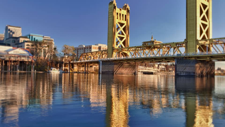 A bridge with a lattice structure spanning over a body of water, with buildings and a tower in the background.