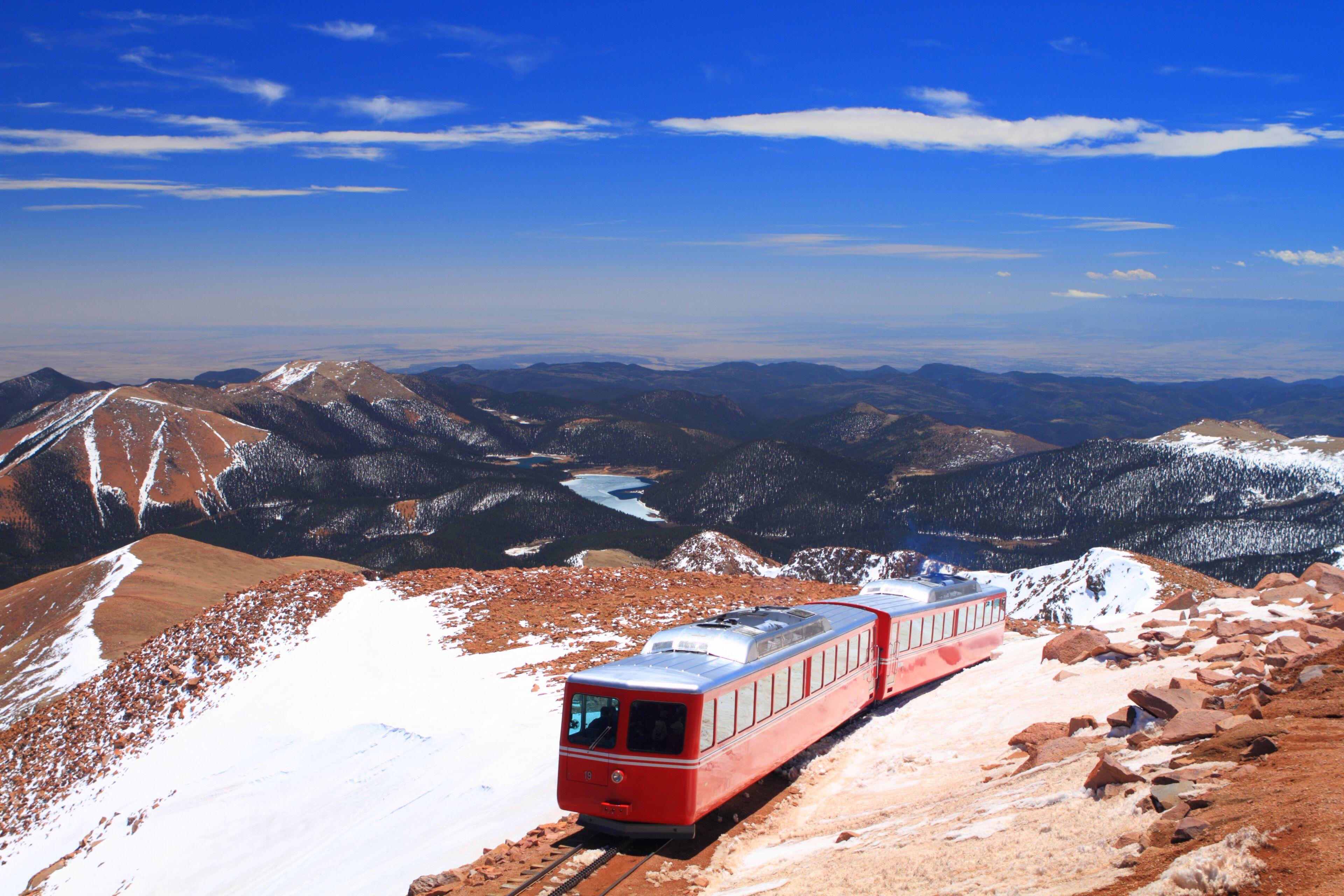 A red train on tracks through a mountainous landscape with snow-covered peaks and a clear blue sky.
