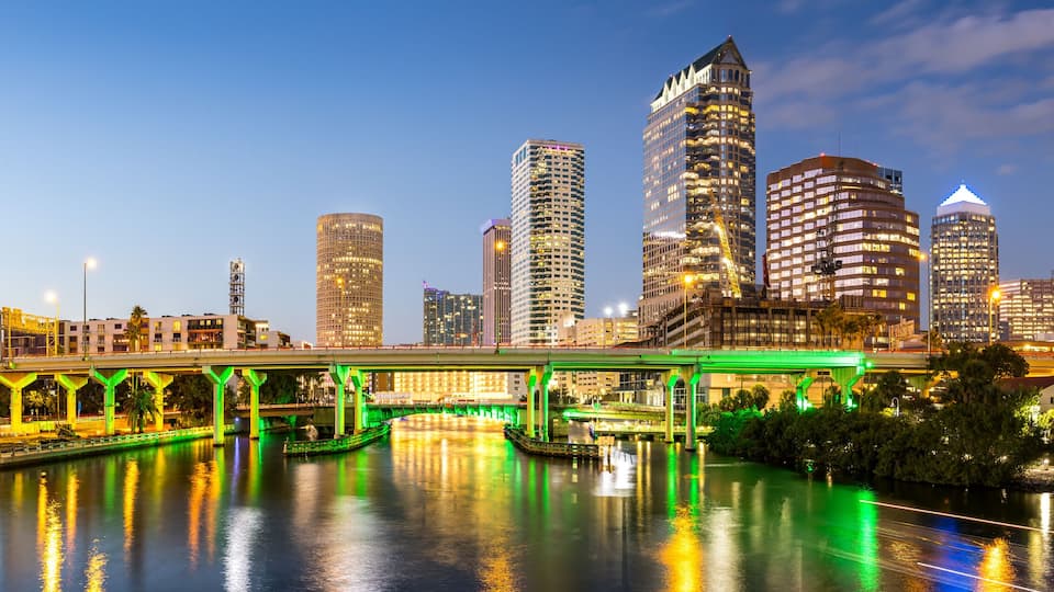 A city skyline at dusk with illuminated buildings and a green-lit bridge over a river.