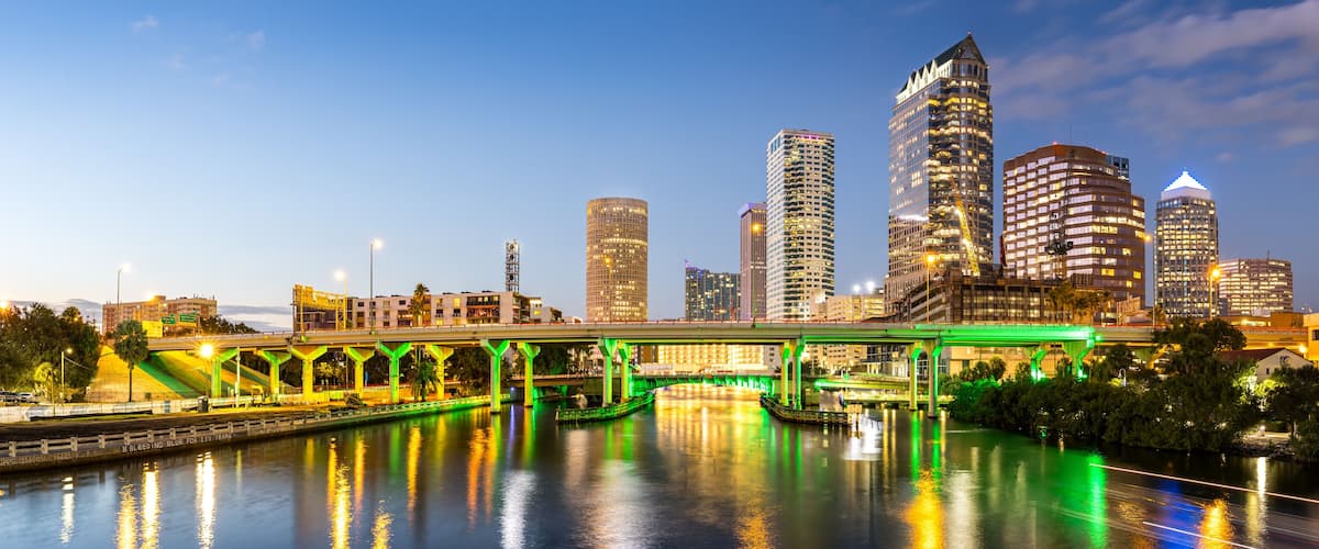 A city skyline at dusk with illuminated buildings and a green-lit bridge over a river.