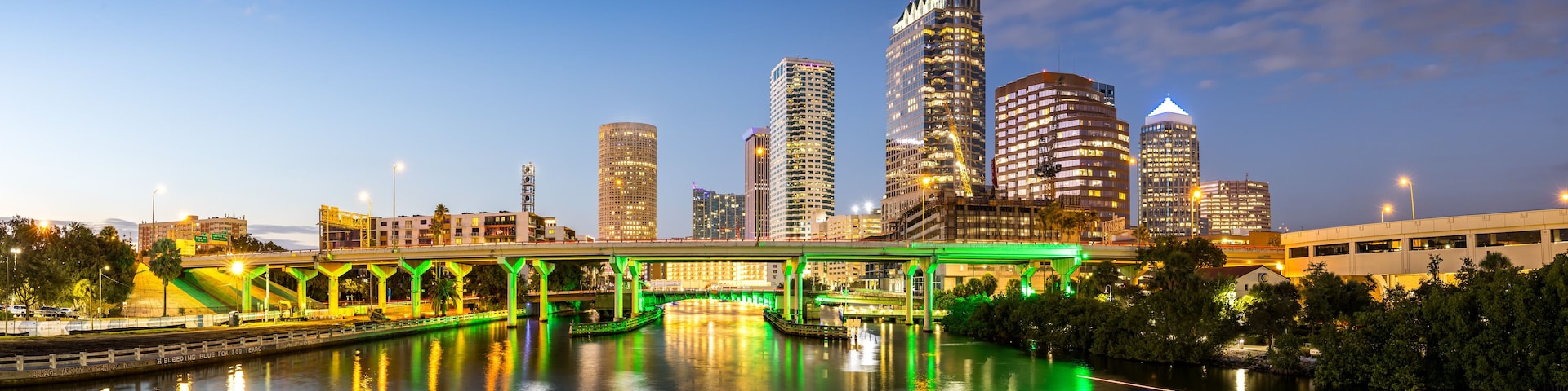 A city skyline at dusk with illuminated buildings and a green-lit bridge over a river.