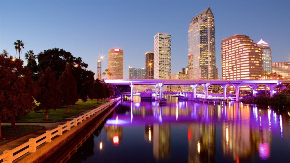 A city skyline with illuminated buildings, a bridge over a river, and a park with trees.