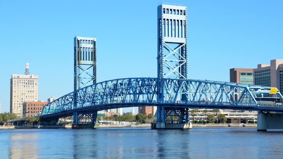 A blue steel bridge over a river with tall buildings in the background.