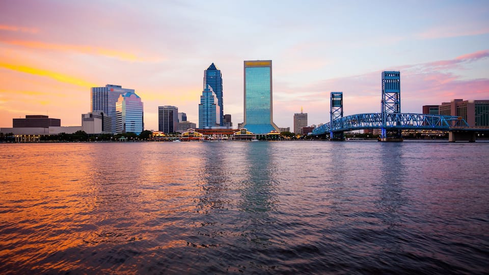 A city skyline with modern buildings and a blue bridge over a river at sunset.