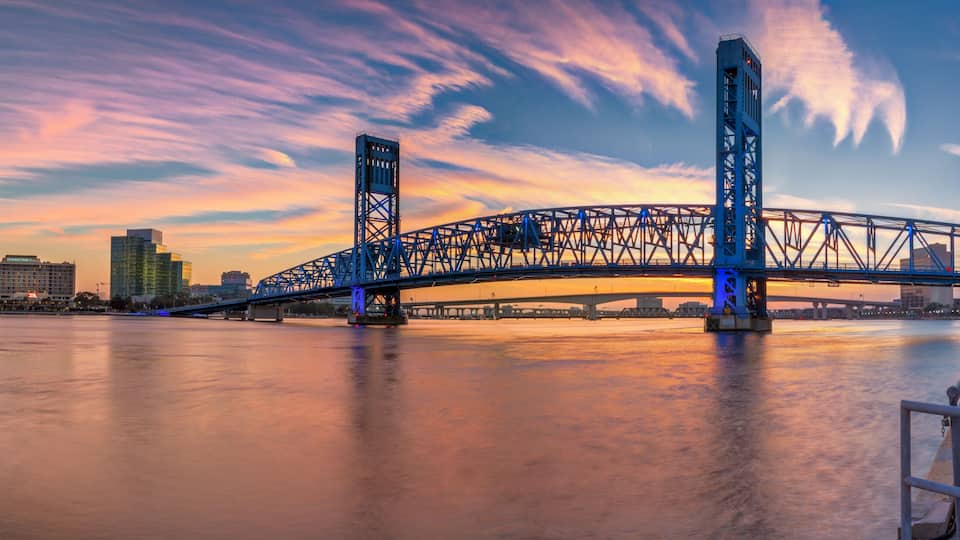 A blue bridge over a river at sunset with a city skyline in the background.