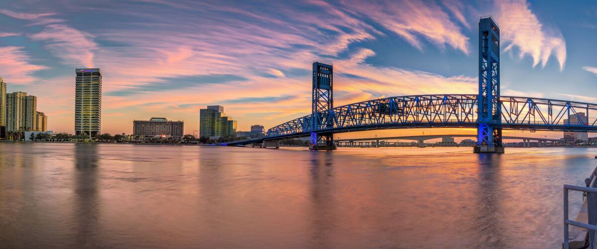 A blue bridge over a river at sunset with a city skyline in the background.