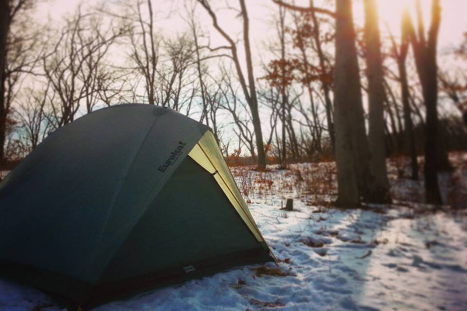 A tent in a snowy forest with bare trees and the sun setting in the background.